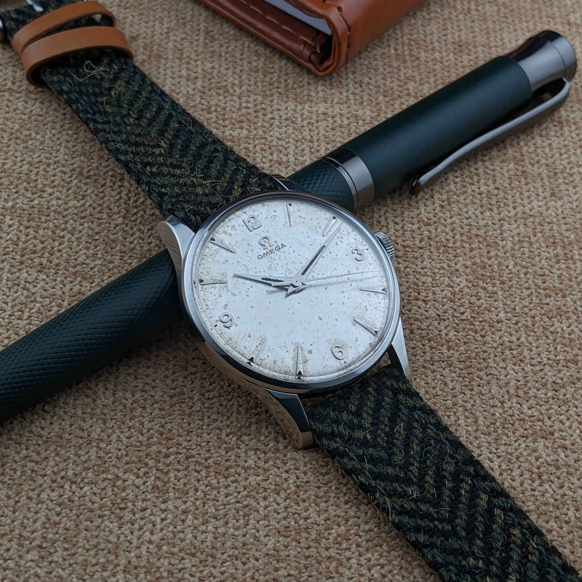 Omega Classic Referenz 2810-2 SC diagonal über zwei dunklen Stiften, mit einem braunen Notizbuch und Armbandschließe im Hintergrund.