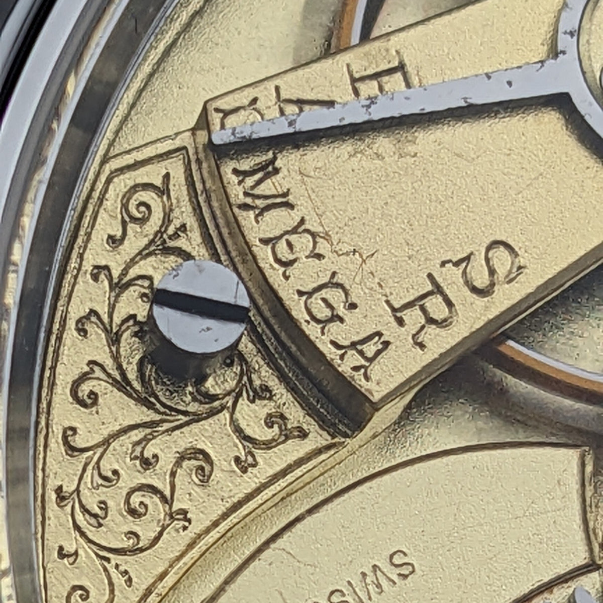 Primo piano del movimento Omega con decorazione floreale incisa, vite a fessura, scritte e parte di un ingranaggio dorato.