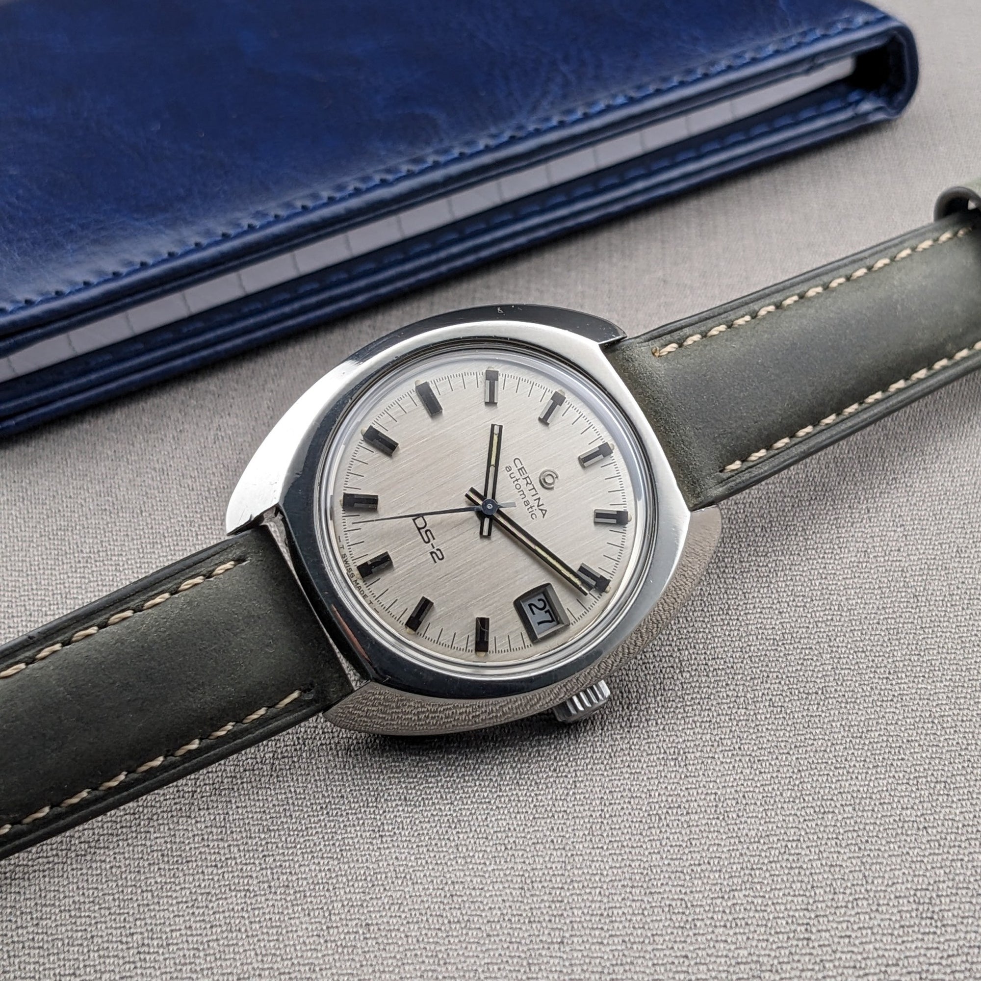 Angled close-up of the Certina DS-2 Automatic reference 5801 300 beside a blue wallet, showing its silver dial and gray-green strap.