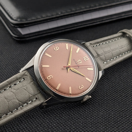 Omega Classic 2760-6 SC mostrato in diagonale con quadrante rosa e cinturino grigio a motivo coccodrillo, accanto a portafoglio nero su tessuto testurizzato.