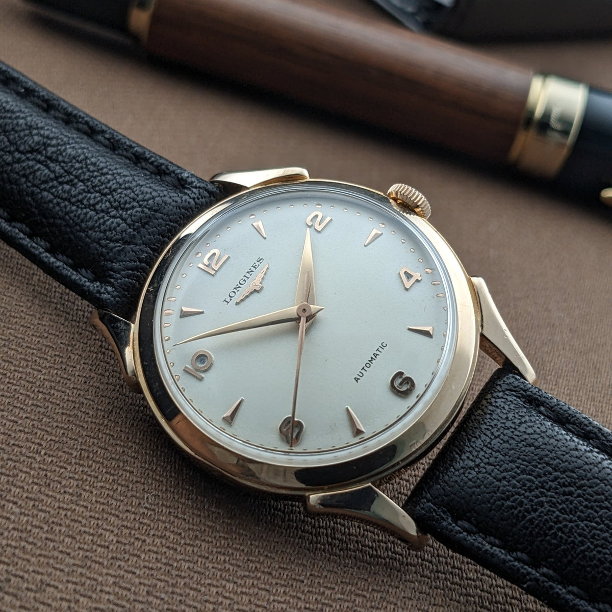 Nahaufnahme des Zifferblatts und goldfarbenen Gehäuses der Longines Automatik Referenz 6058 auf braunem Stoff, mit einem Stift dahinter.
