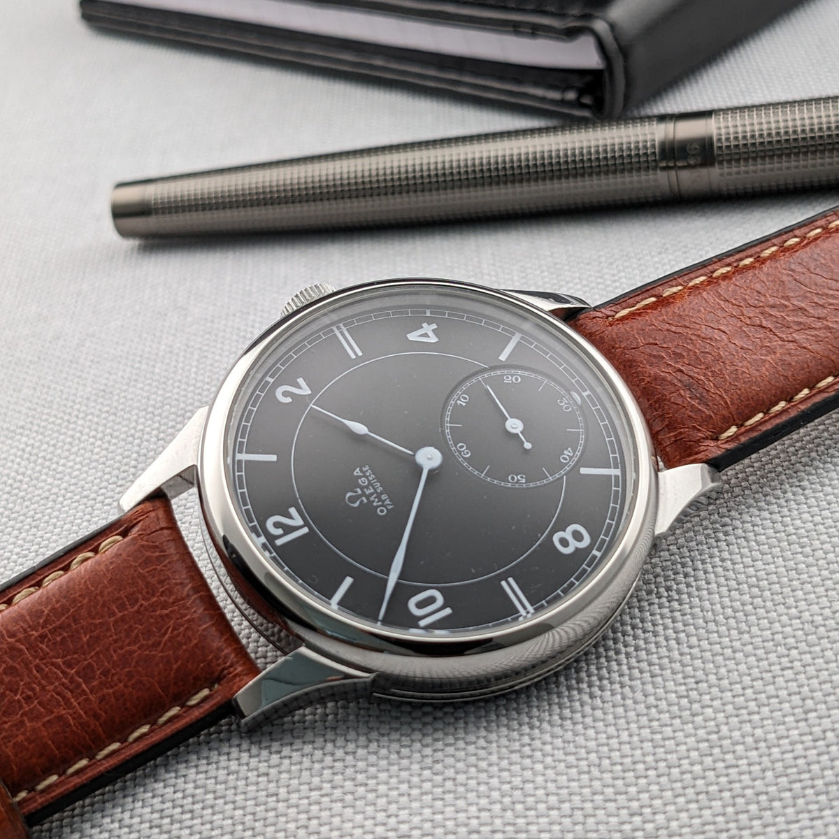 Omega-Armbanduhr schräg auf einer strukturierten Oberfläche, mit braunem Lederarmband, dunklem Zifferblatt, silbernem Stift oben und schwarzem Etui im