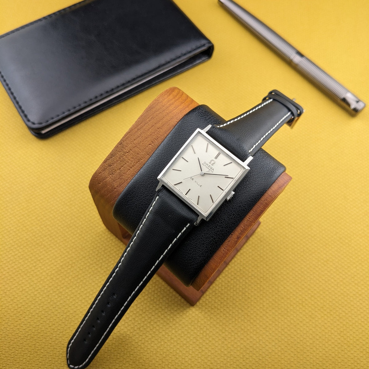 Omega De Ville Automatik 161.014 auf einem Uhrenkissen, mit ausgezogenem schwarzen Armband neben schwarzem Gehäuse und silbernem Stift.
