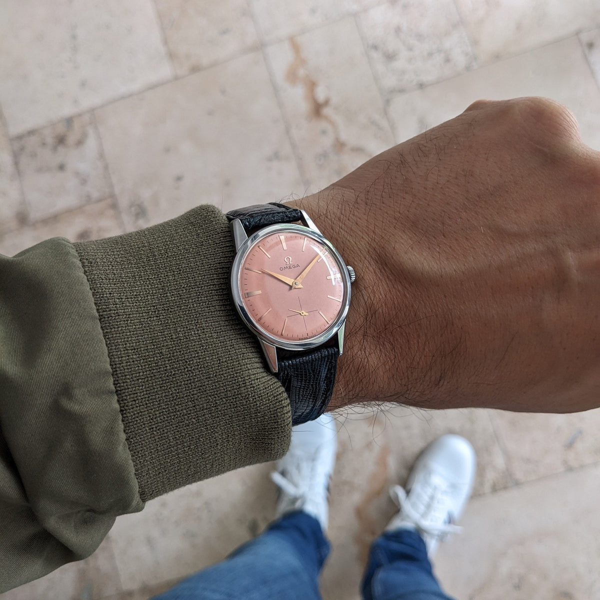 Omega Classic referenza 14391-1 indossato al polso con cinturino nero, mostrato su una manica oliva con pavimento piastrellato e scarpe bianche sotto.
