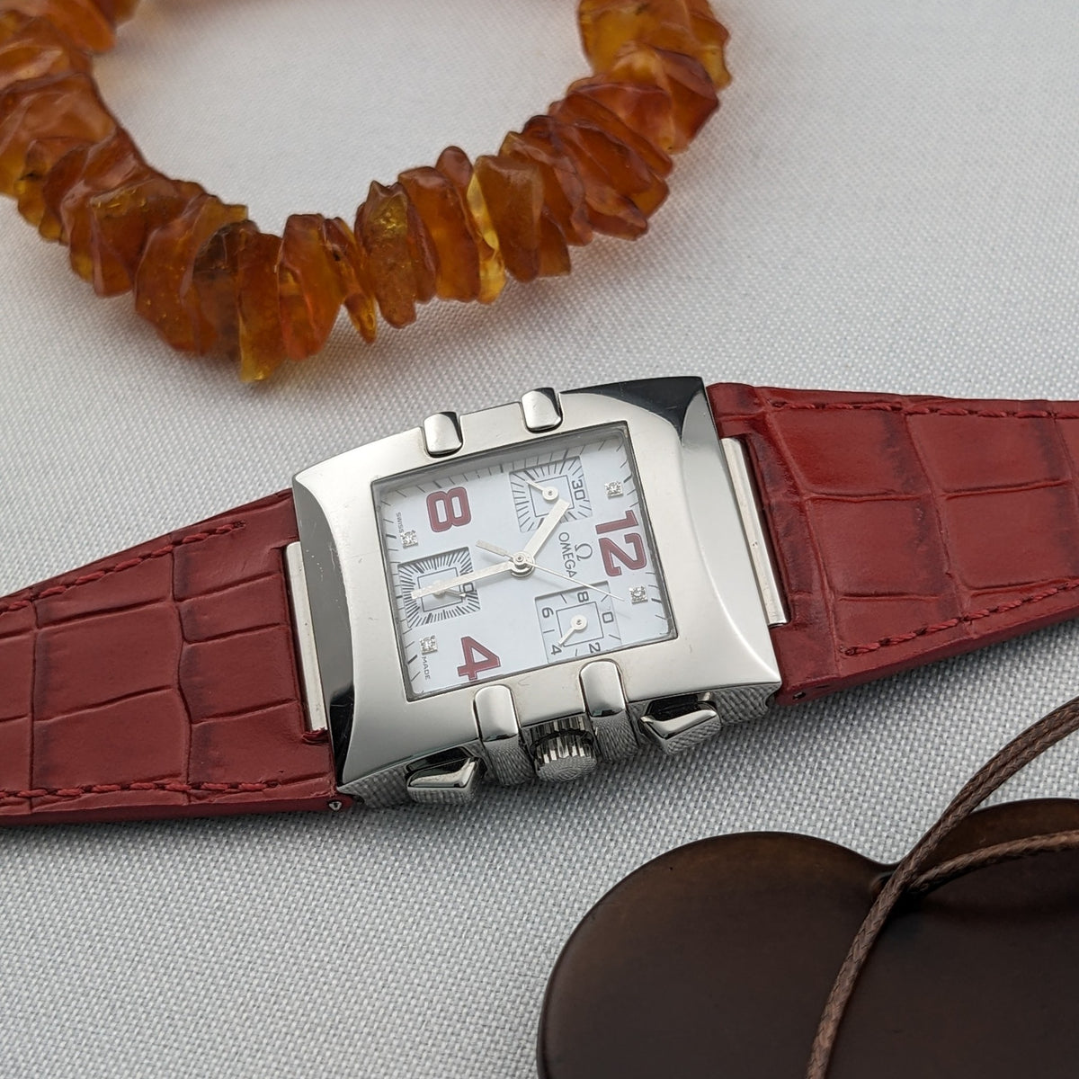 Omega Constellation Quadra 1841.73.31 shown horizontally with a white dial, red strap, amber bead strand, and brown pendant.