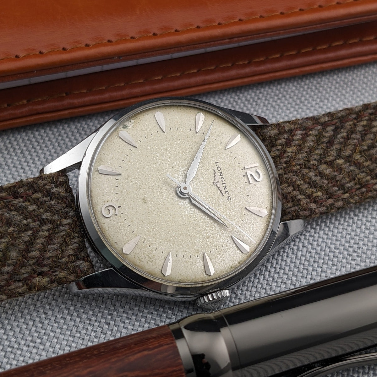 Longines Classic reference 8135-5 lies diagonally beside a leather wallet and dark wood pen, with its cream dial and woven strap prominent.