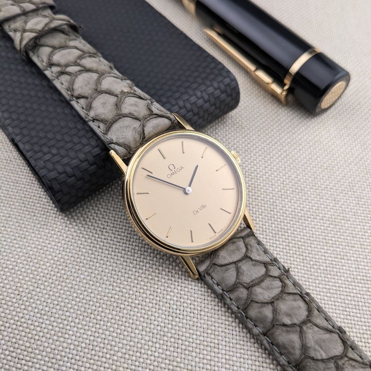 Omega De Ville reference 111 0156 angled across a dark case, showing its cream dial, gold-tone case, gray strap, and nearby pen.