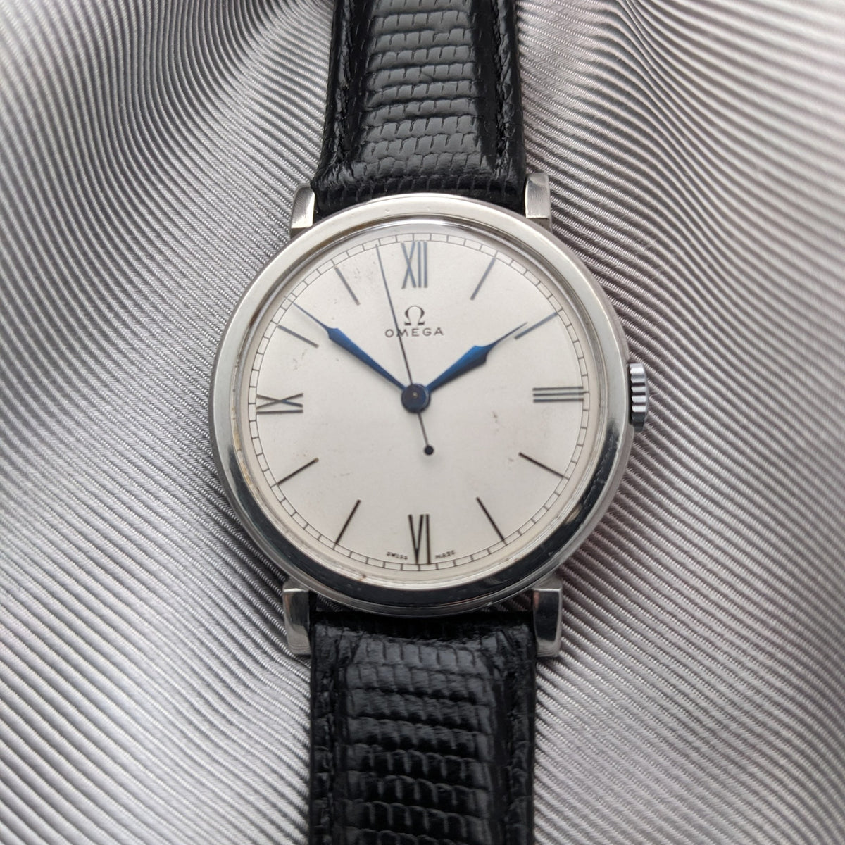 Front-facing Omega watch on gray fabric, featuring a light dial, Roman numerals, blue hands and a black strap.