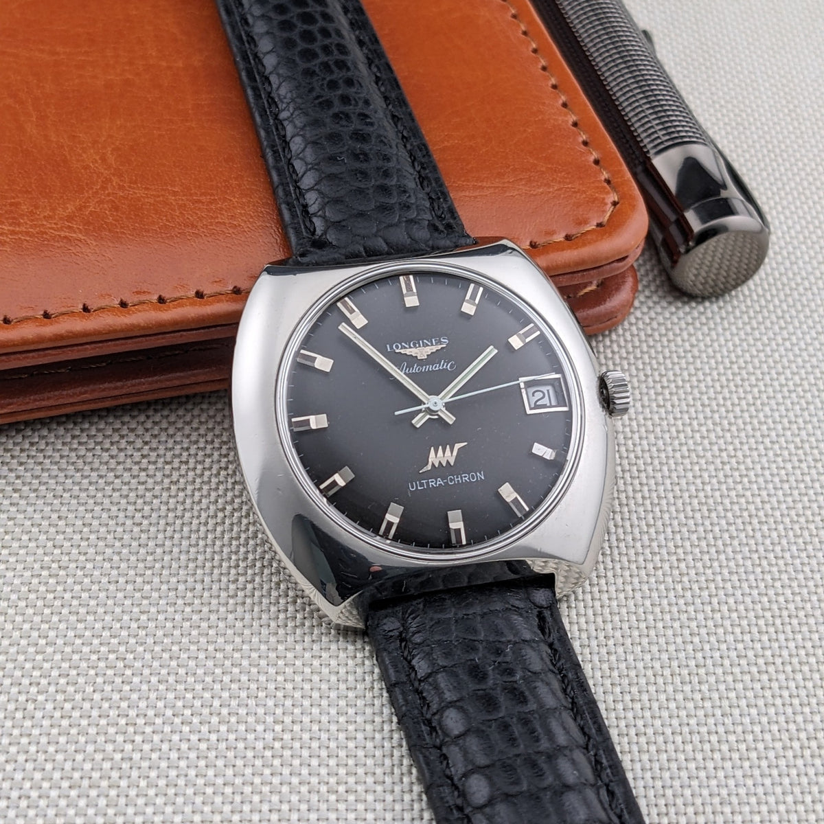 Longines Ultrachron reference 8429 shown upright against a brown wallet, with dark dial, date window and black patterned leather strap.