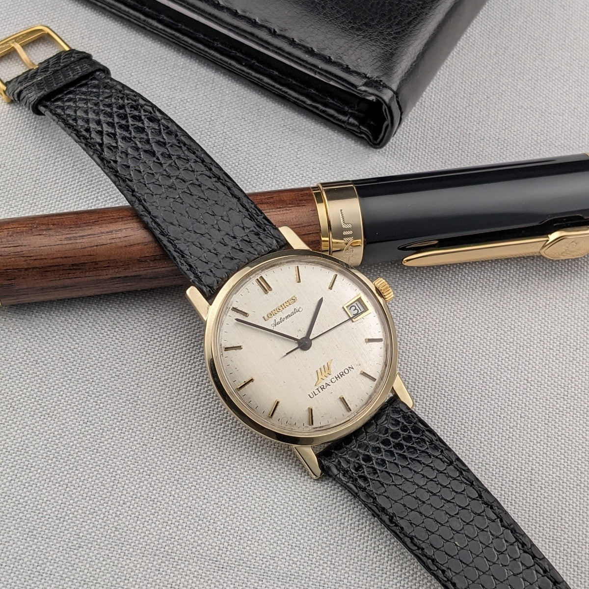 Longines Ultra-Chron reference 69-556AC arranged diagonally beside a black case and wood-and-black pen on light fabric.
