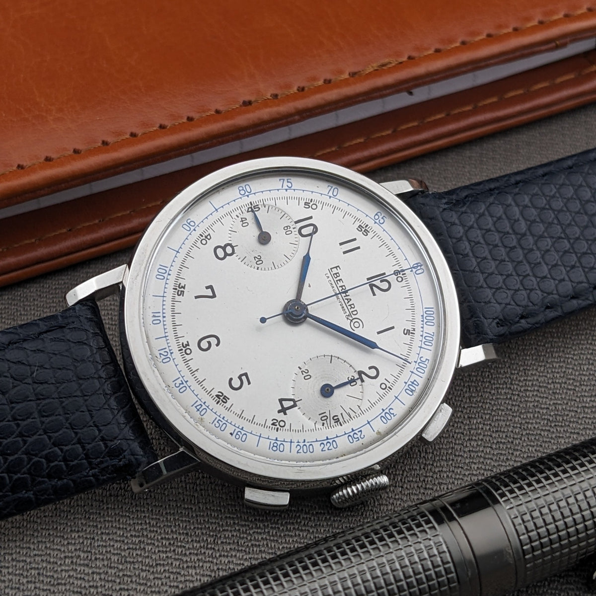 Angled Eberhard & Co. Pre-Extra Fort dial with cream face, blue outer scales, black numerals and blue strap, framed by a wallet and pen.