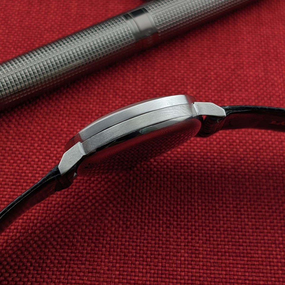 Low side view of the Longines Calatrava reference 20757 case and strap, with a patterned silver pen crossing the upper background.