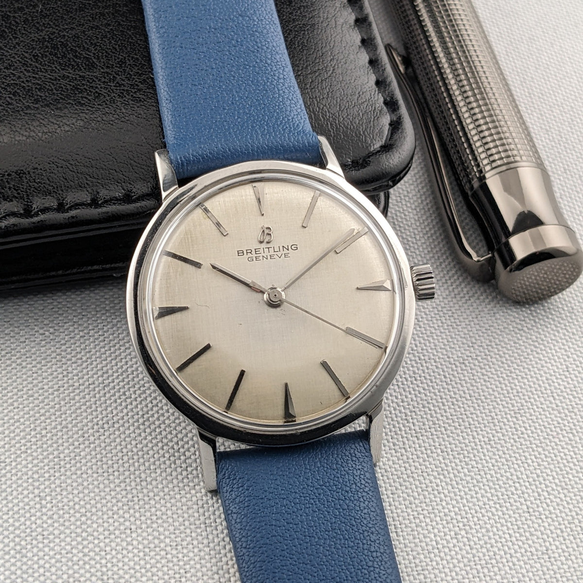 Breitling Automatic watch at a three-quarter angle with cream dial and blue strap, beside a black case and silver pen.