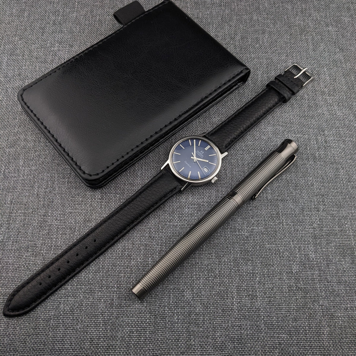 Omega Geneve Automatic reference 166.0163 pictured with its black leather strap, black rectangular case and silver textured writing instrument.