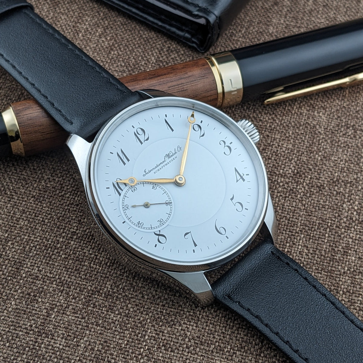 IWC Schaffhausen घड़ी का तीन-चौथाई दृश्य, काले पट्टा, पॉलिश केस, सफेद डायल और सुनहरे हाथ के साथ लकड़ी के पेन के बगल में।