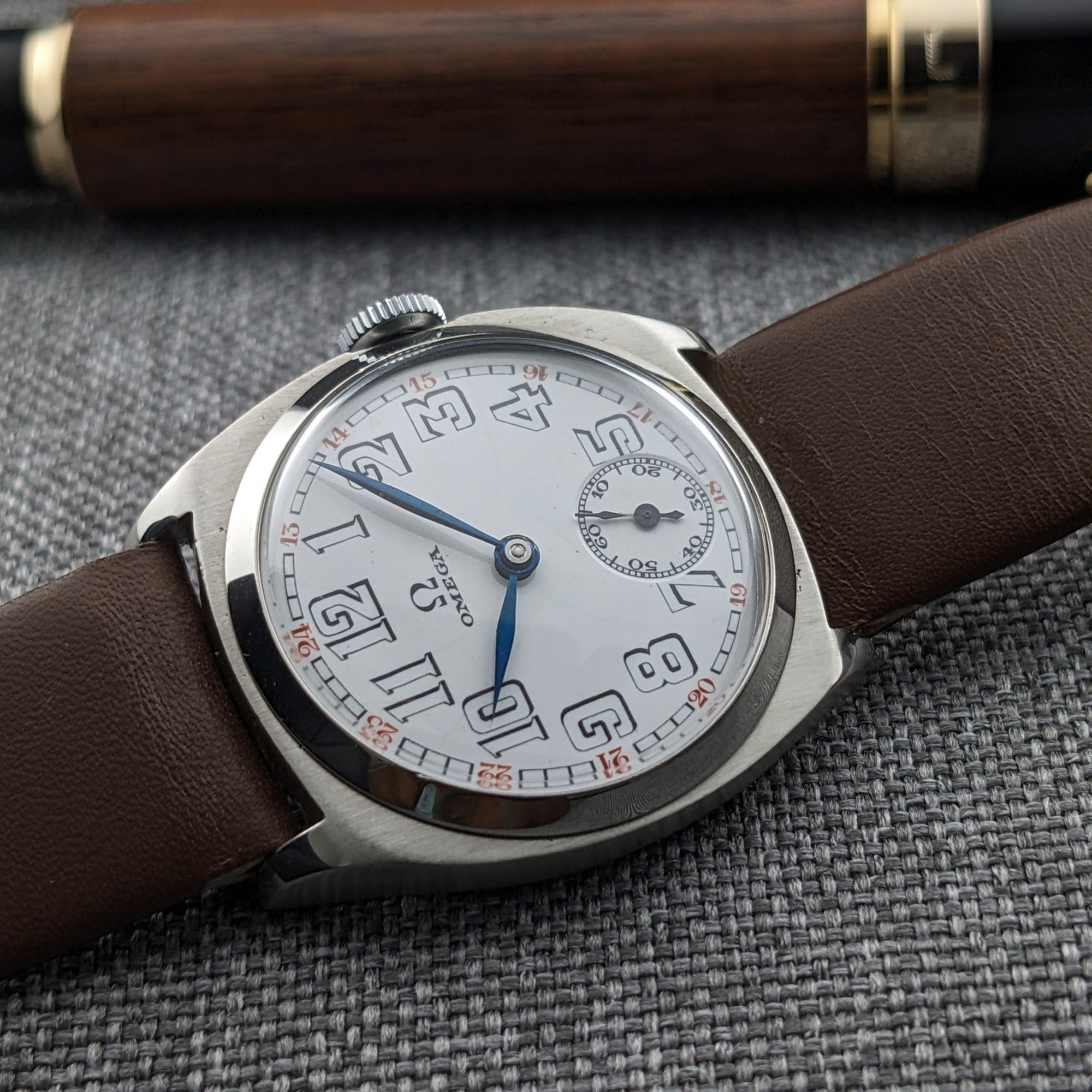 Angled Omega watch composition on gray fabric, showing the white dial, blue hands, Arabic numerals, brown strap, and a wood-and-black pen behind it.