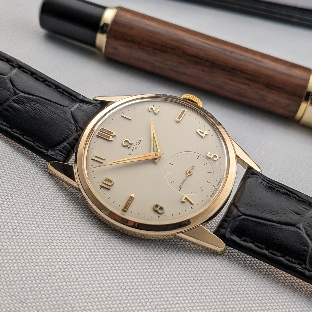 Omega Classic reference 14391-1 lying diagonally on fabric, with its gold-tone dial and black strap framed by a wooden writing instrument.