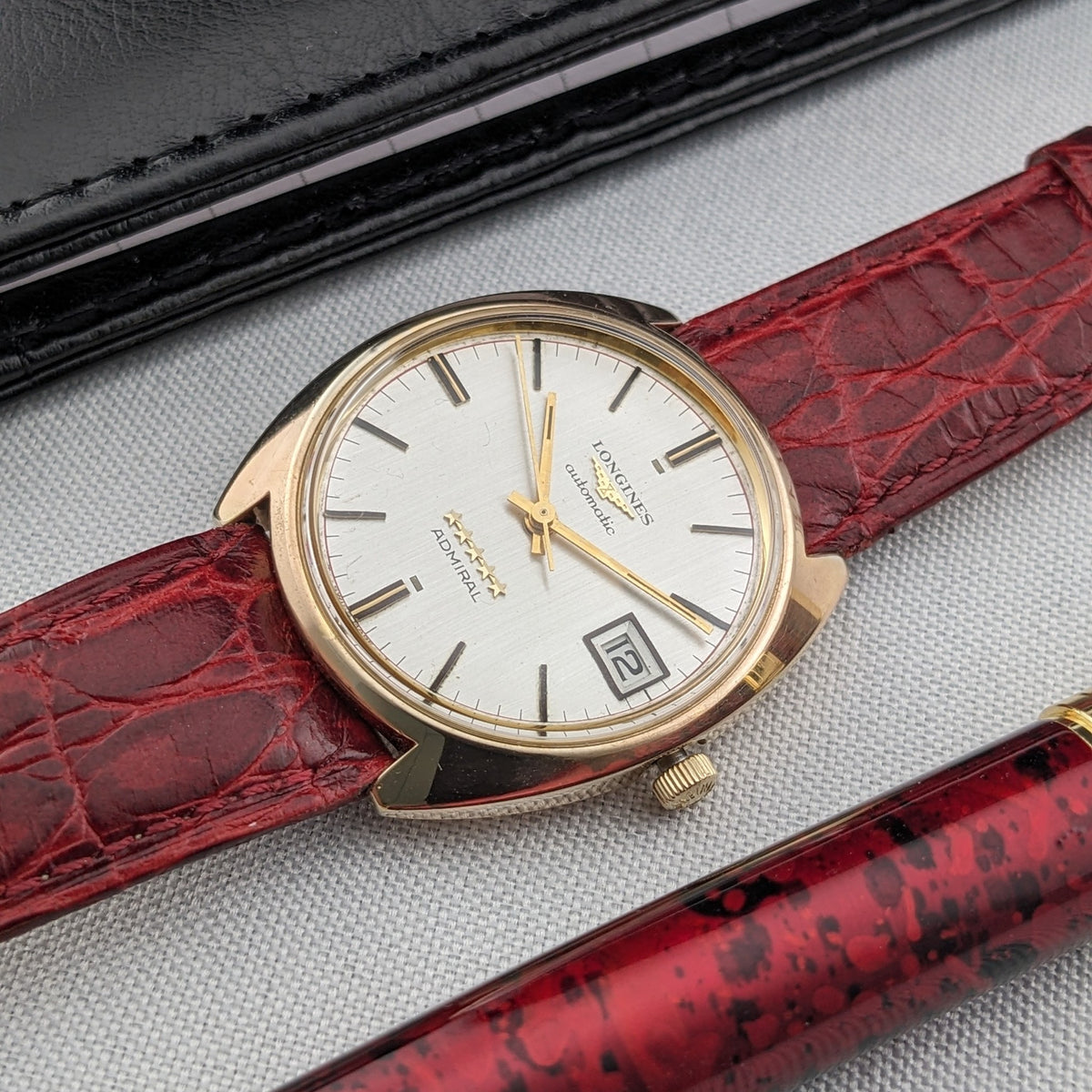 Schrägansicht der Longines Admiral Referenz 8343-1 auf einer strukturierten Oberfläche, mit silbernem Zifferblatt, Datumsfenster, Armband und Stift.