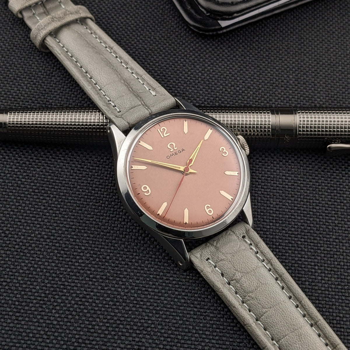 Omega Classic Referenz 2760-6 SC über einem strukturierten Metallstift, zeigt das rosafarbene Zifferblatt, graues Armband und schwarzen Stoffhintergrund.