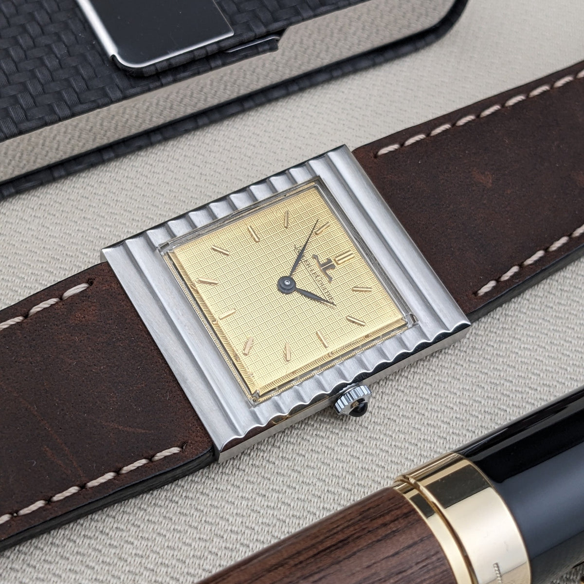 Jaeger-LeCoultre Vogue Tank Ref. 9120 diagonal gezeigt mit ihrem goldfarbenen Zifferblatt, gerippten Gehäusekanten, braunem Armband und einem nahen Stift.
