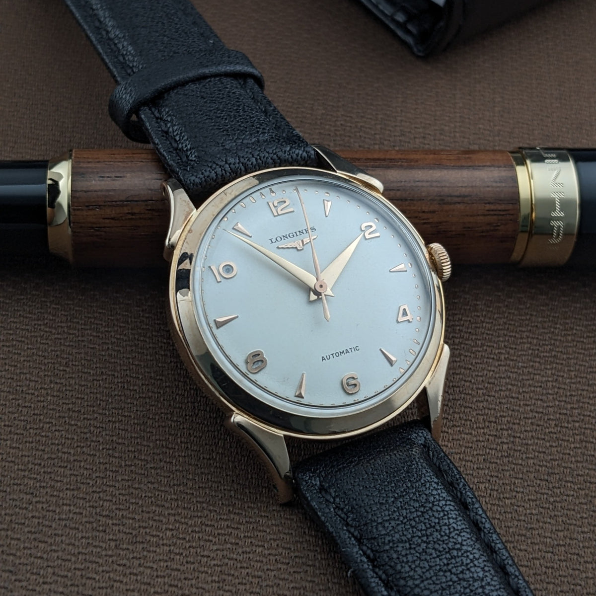 Longines Automatik Referenz 6058 schräg auf braunem Stoff, über einem Holzstift positioniert, zeigt ihr helles Zifferblatt und schwarzes Armband.