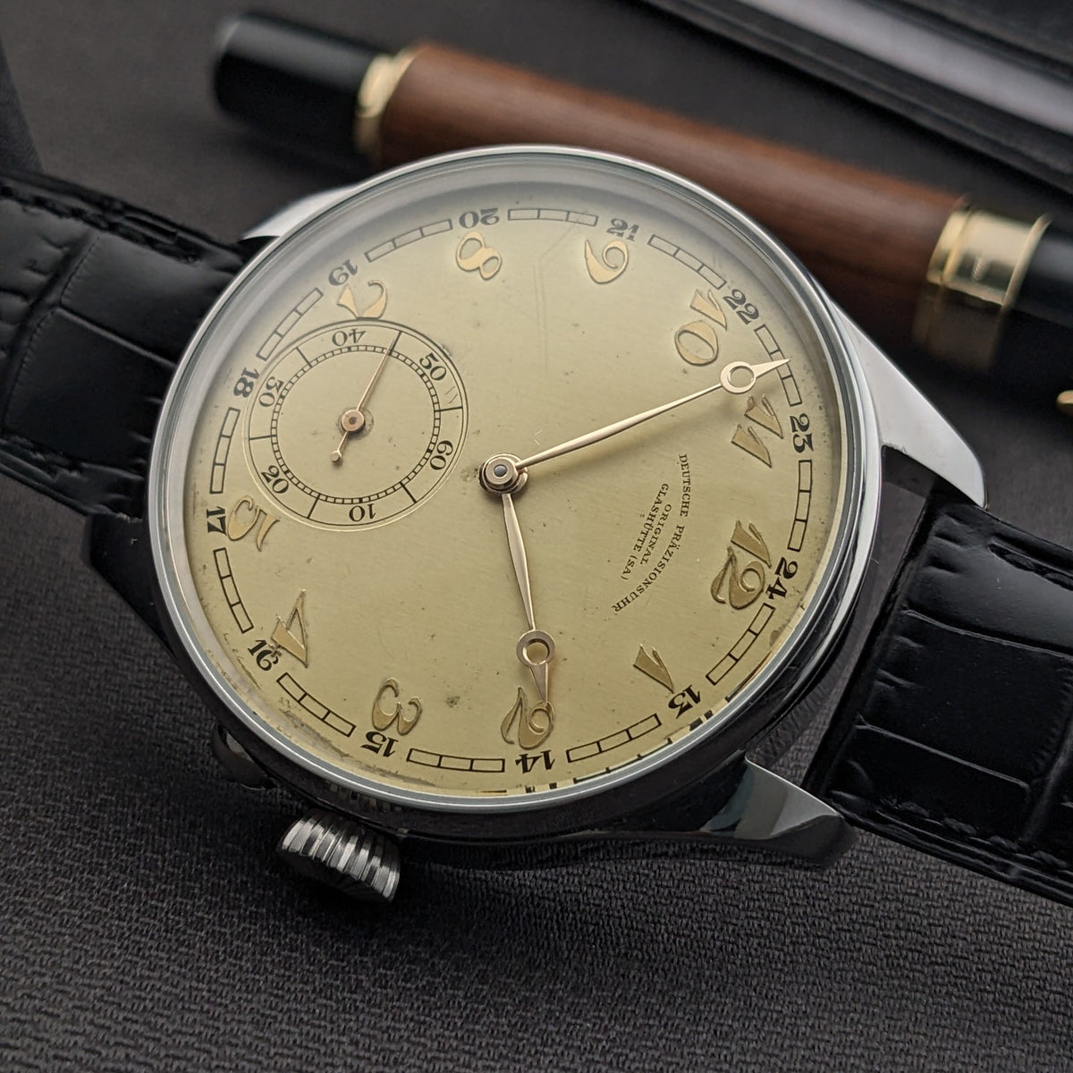 Schrägansicht der Deutsche Präzisions-Uhrenfabrik Glashütte Uhr auf dunklem Stoff, zeigt das cremefarbene Zifferblatt, kleine Sekunde, Krone und schwarzes
