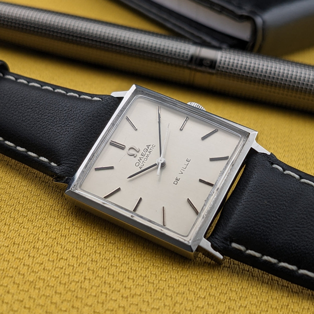 Nahaufnahme der Omega De Ville Automatik 161.014, hebt helles Zifferblatt, quadratisches Gehäuse, schwarzes Armband, Stift und senffarbenen Hintergrund hervor.