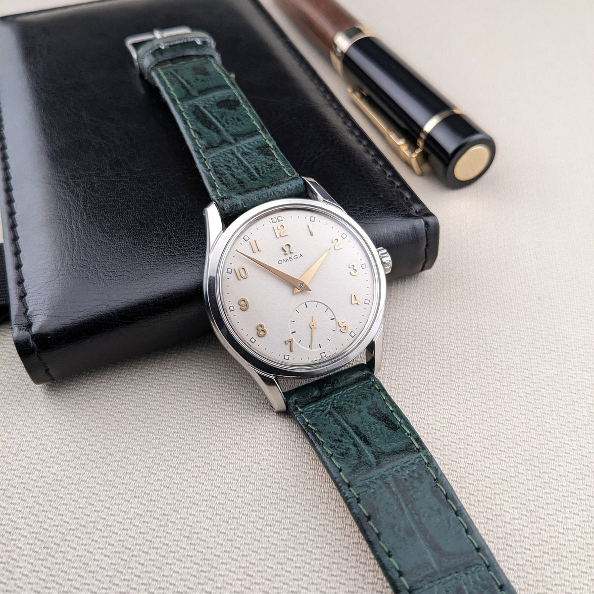 Vista a tre quarti dell'OMEGA Classic referenza 2639-11 su una custodia in pelle nera, con cinturino verde, quadrante argentato e penna vicina.