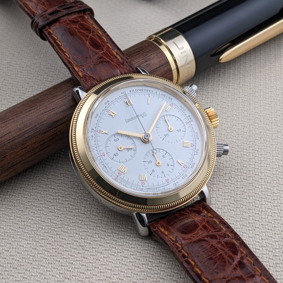 Eberhard & Co.. Godron Chrono reference 32031 rests over a wooden and black fountain pen, with its white dial and brown strap in view.