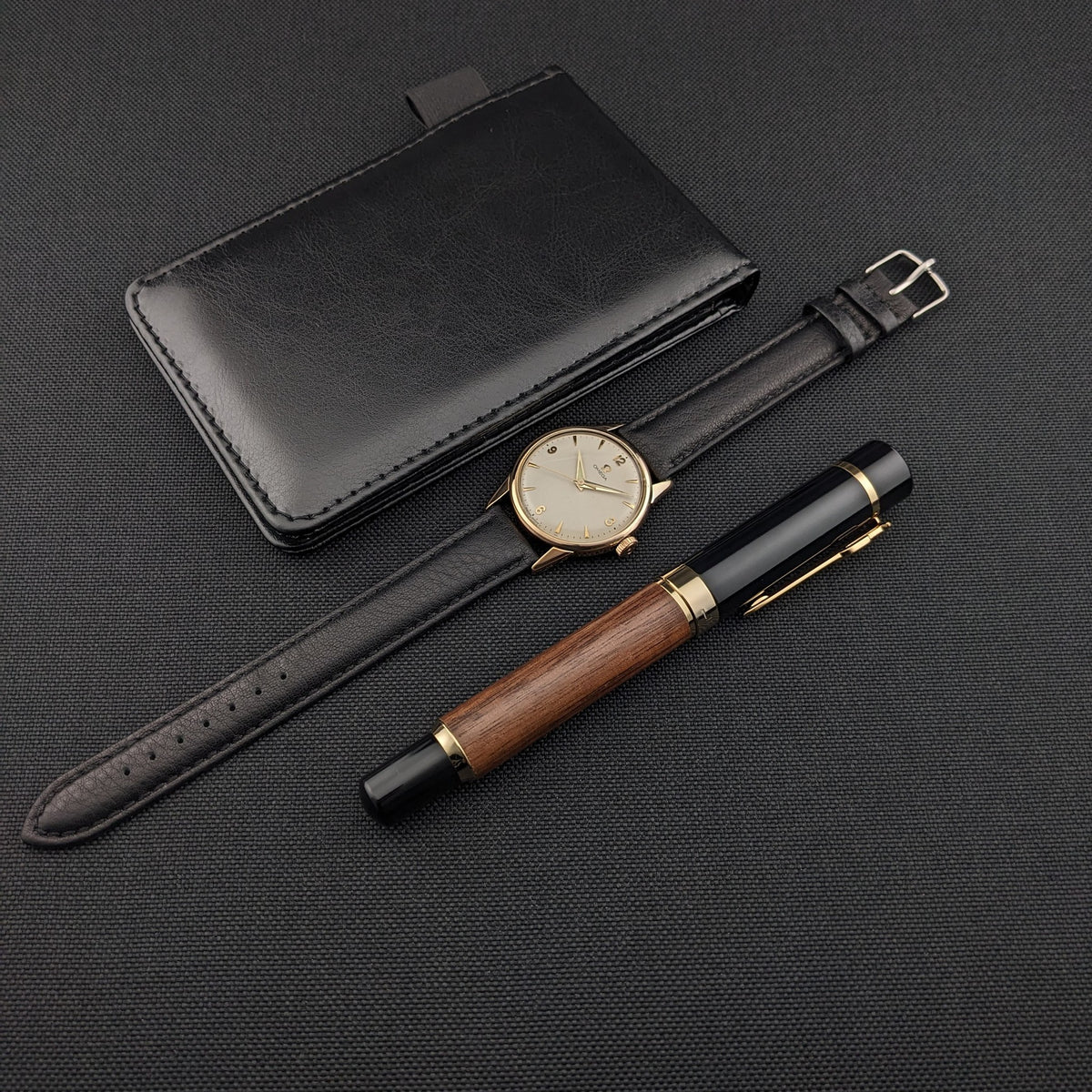 Omega Classic référence 14392-61-SC avec son bracelet en cuir noir posé à côté d'un étui noir et d'un stylo en bois et noir.