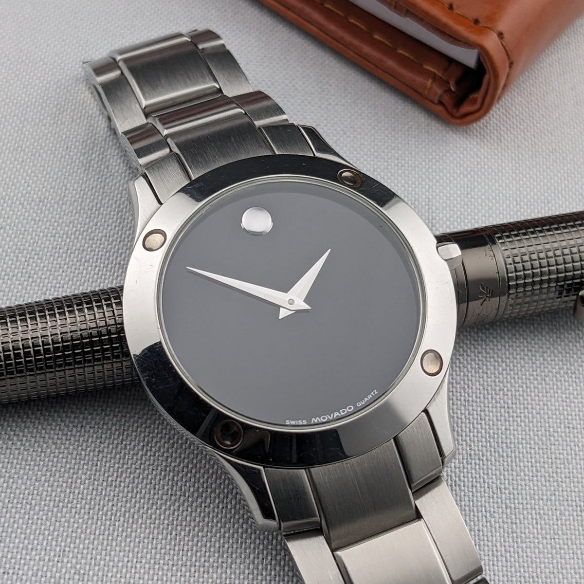 Montre Movado placée sur un stylo texturé foncé, montrant son cadran noir, aiguilles argentées, lunette polie et bracelet en acier.