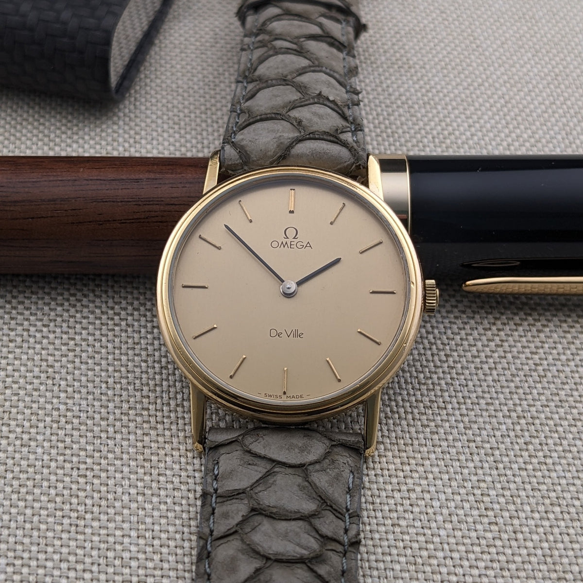 Omega De Ville reference 111 0156 centered over a wood-and-black pen, with cream dial, baton markers, and gray crocodile-pattern strap.
