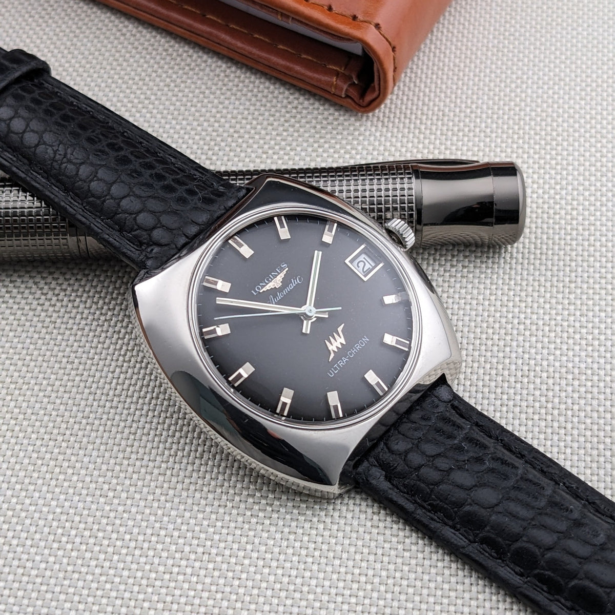 Longines Ultrachron reference 8429 displayed over a textured silver pen, showing its dark dial, date window, black strap and brown wallet.