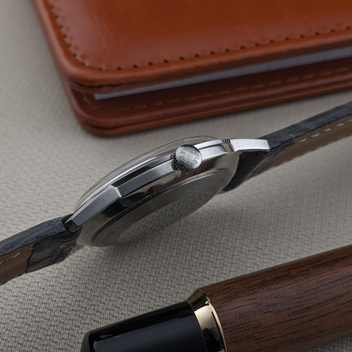 Side profile of the Longines Ultra-Chron 7827 showing the crown, case edge, lugs, and black strap against a leather case and pen.