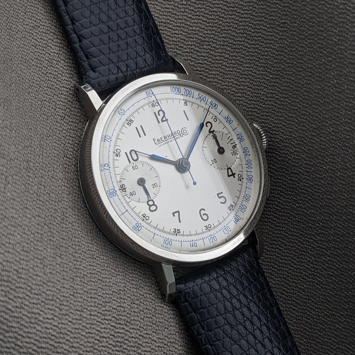Eberhard & Co.. Pre-Extra Fort shown at an angle on gray fabric, highlighting the cream dial, two subdials, crown and dark blue strap.