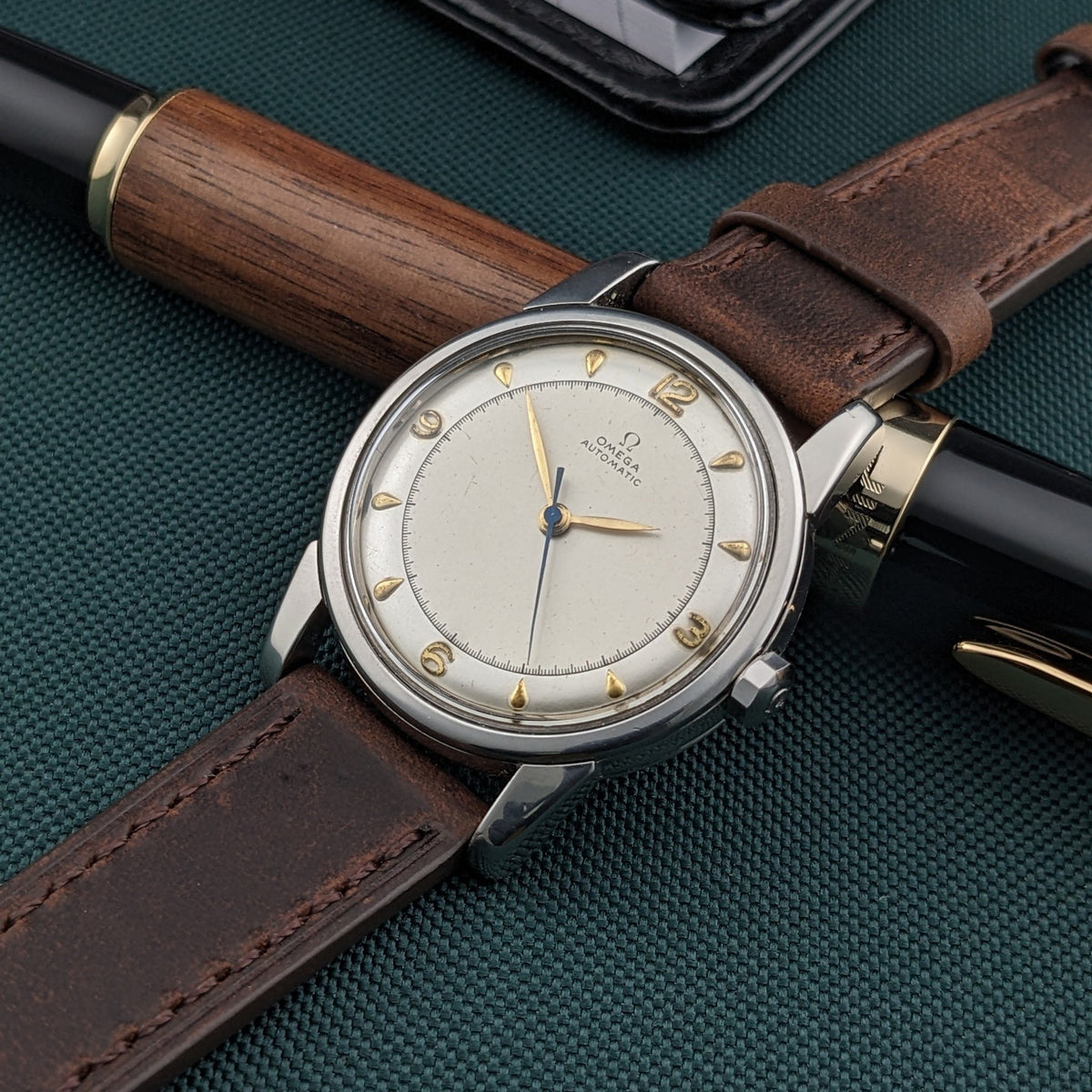 Three-quarter view of the Omega Automatic Bumper reference 2767-4 SC with cream dial and brown strap, placed beside a wood-and-black pen.