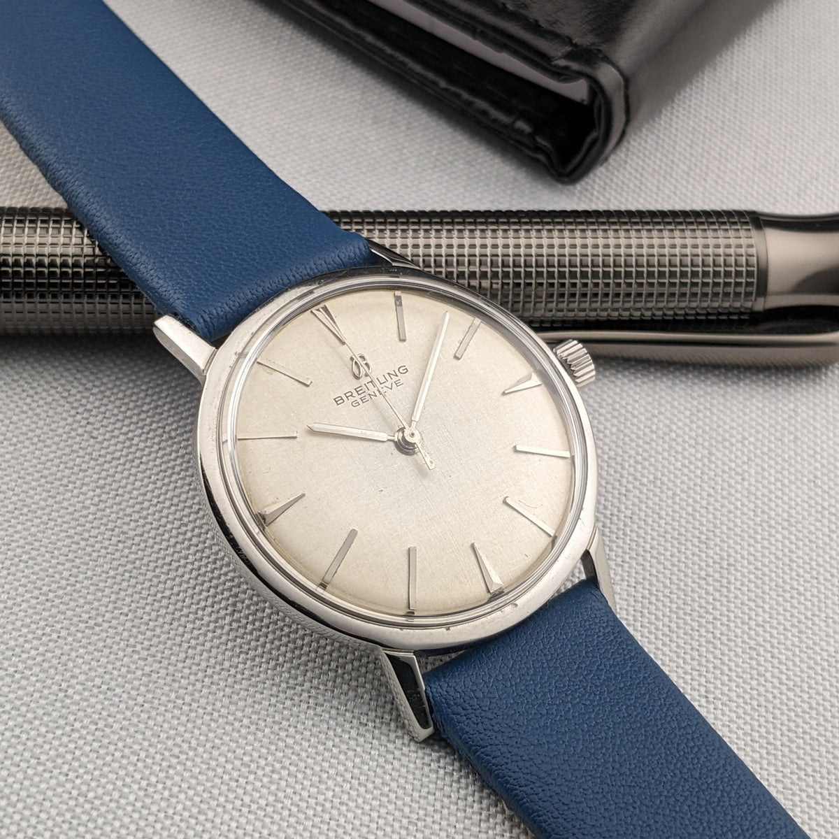 Angled front view of the Breitling Automatic watch with cream dial and blue strap laid across a silver textured pen.