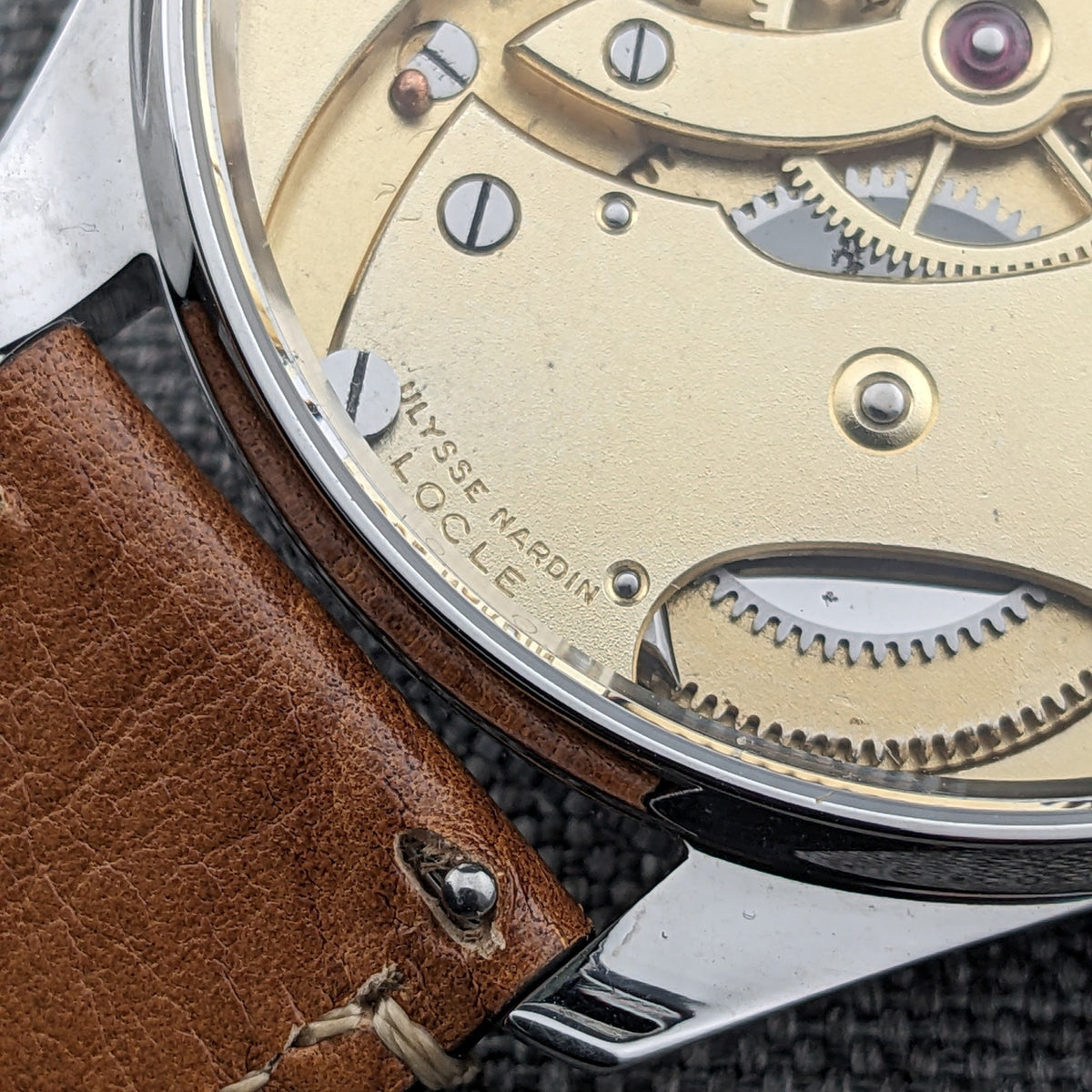 Ulysse Nardin मूवमेंट का विस्तृत दृश्य केस एज के पास, जिसमें दृश्य गियर्स, उत्कीर्ण अक्षर और भूरा पट्टा कनेक्शन दिखता है।