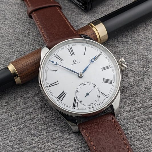 Omega wristwatch photographed beside a wood-and-black pen, showing its white Roman-numeral dial, blue hands and brown leather strap.