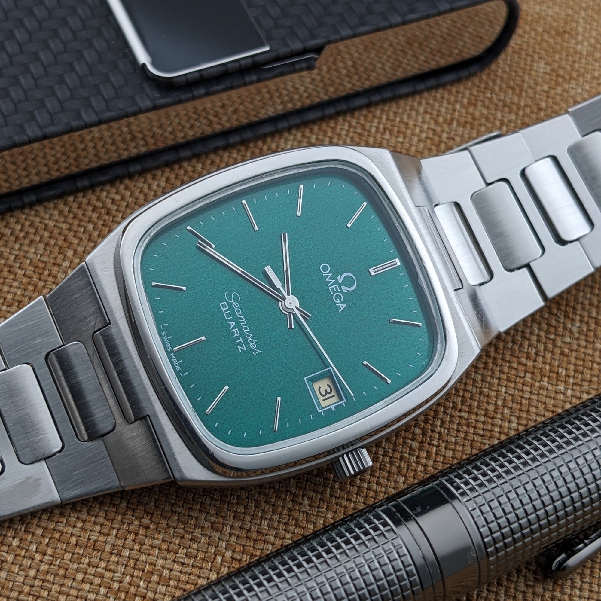 Close-up of the Omega Seamaster reference 196.0166 teal dial and date window, with bracelet links and a textured black pen nearby.