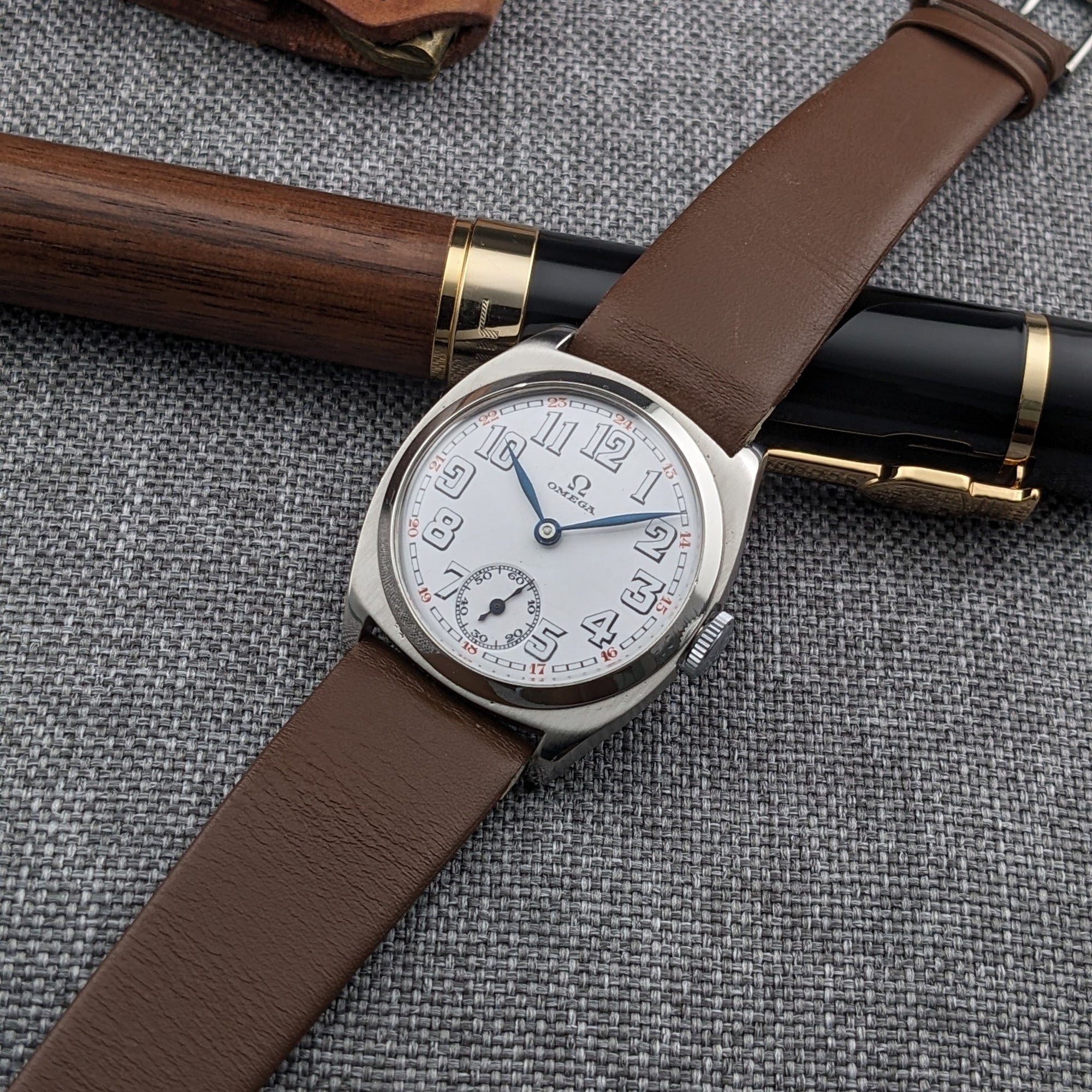 Omega watch with brown leather strap placed diagonally over a black-and-wood pen on gray woven fabric.