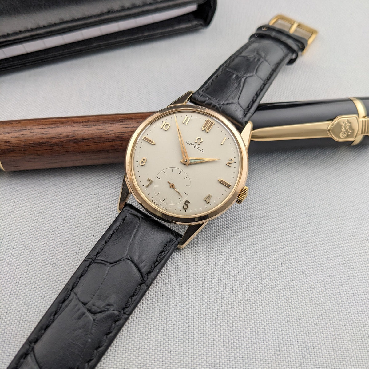 Omega Classic reference 14391-1 laid across a wooden pen and black case, showing the full black strap, gold-tone case and light dial.