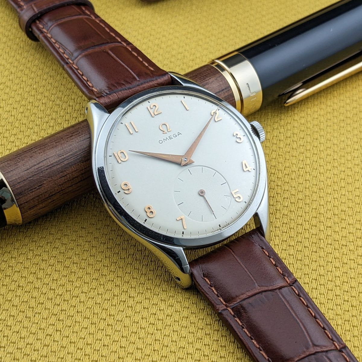 Omega Jumbo Referenz 2505-10 diagonal über einem Holz-Schreibgerät platziert, mit sichtbarem hellem Zifferblatt und braunem Armband.