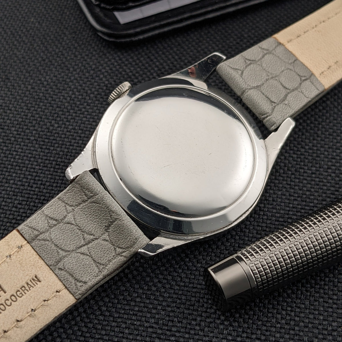 Rückansicht der Omega Classic 2760-6 SC mit einfachem rundem Gehäuseboden, grauem Armband und strukturiertem Metallstift auf schwarzem Stoff.