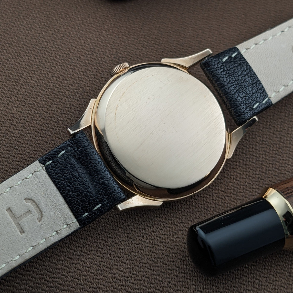 Rückseite der Longines Automatik Referenz 6058 mit glattem rundem Gehäuseboden, schwarzem Lederarmband und einem schwarz-goldenen Stift in der Nähe.