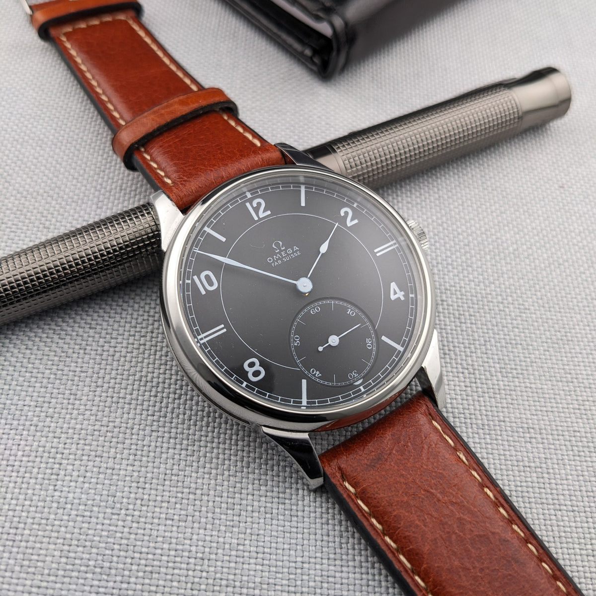 Omega-Armbanduhr diagonal über einem silbernen strukturierten Stift, zeigt ihr dunkles Zifferblatt, poliertes Gehäuse und braunes Lederarmband.