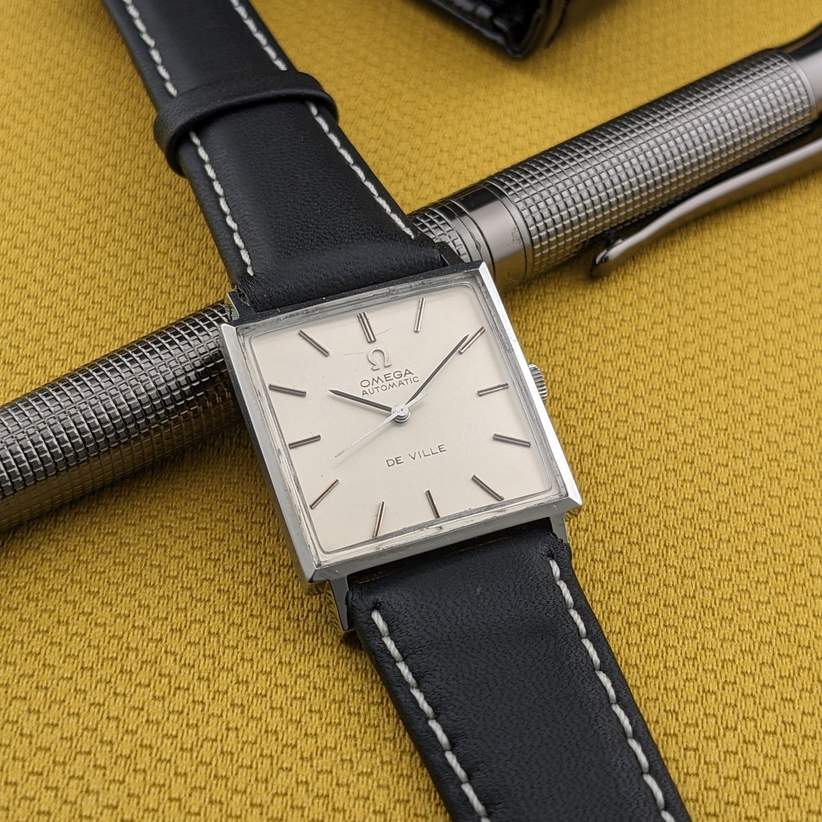 Omega De Ville Automatik 161.014 auf einem silbernen Stift, zeigt helles quadratisches Zifferblatt und schwarzes Armband auf senffarbenem Stoff.
