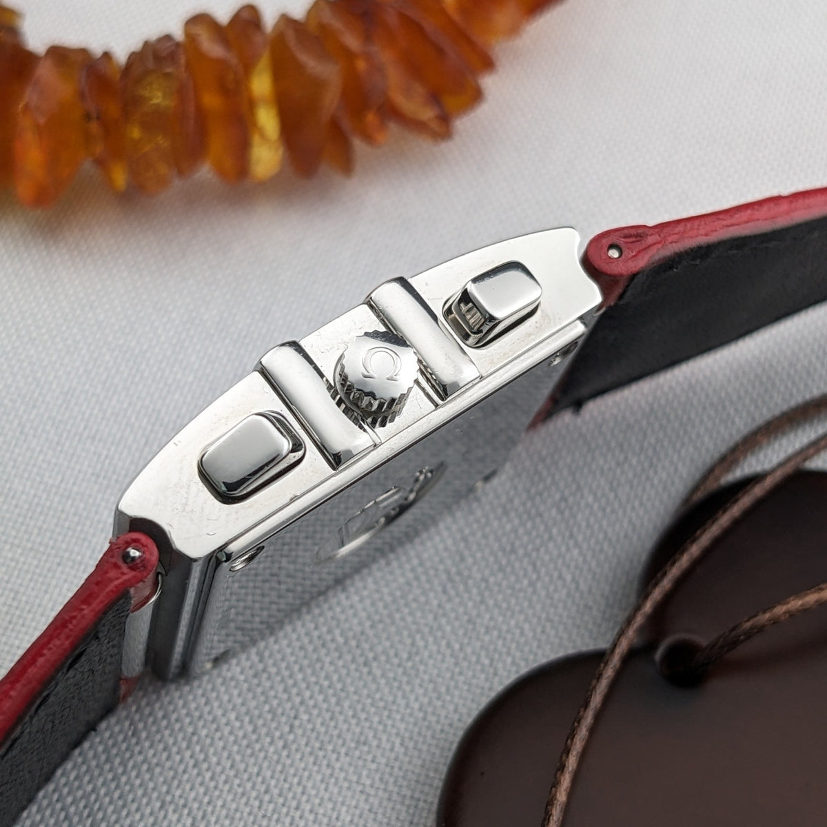 Side view of the Omega Constellation Quadra 1841.73.31 showing its crown, two rectangular side pushers, case edge, and red strap ends.