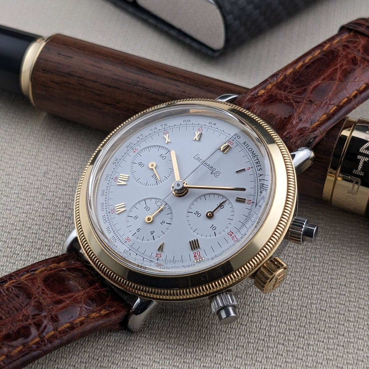 Eberhard & Co.. Godron Chrono reference 32031 shown close-up beside a wooden fountain pen, with gold-tone bezel and three subdials visible.