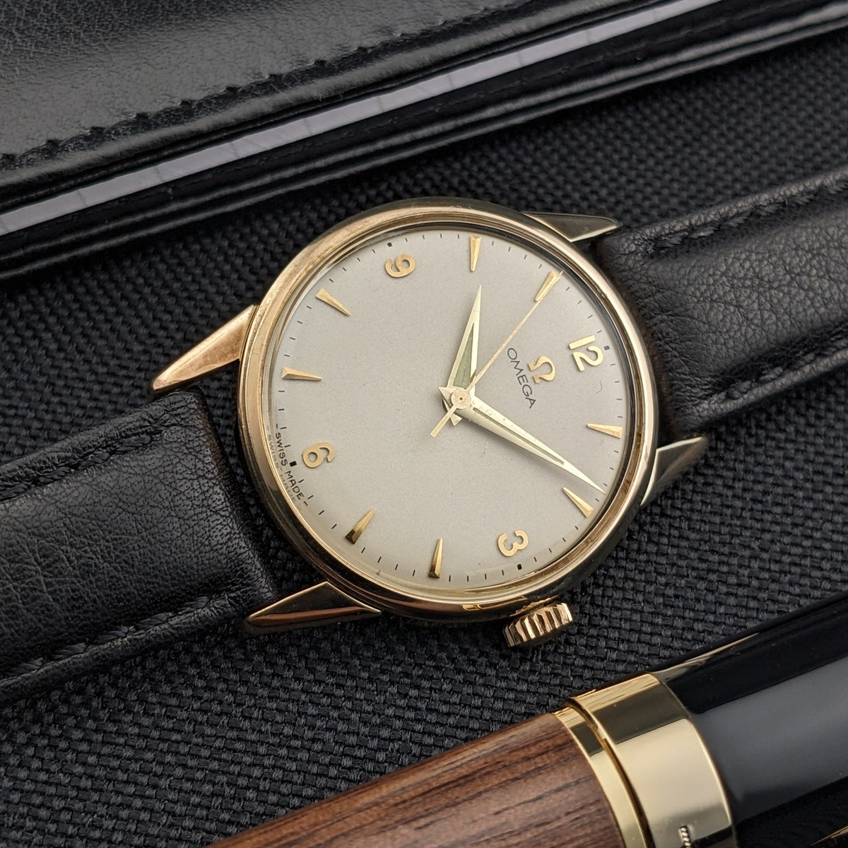 Omega Classic référence 14392-61-SC inclinée avec cadran crème et bracelet noir, accompagnée d'un stylo en bois et noir au premier plan.